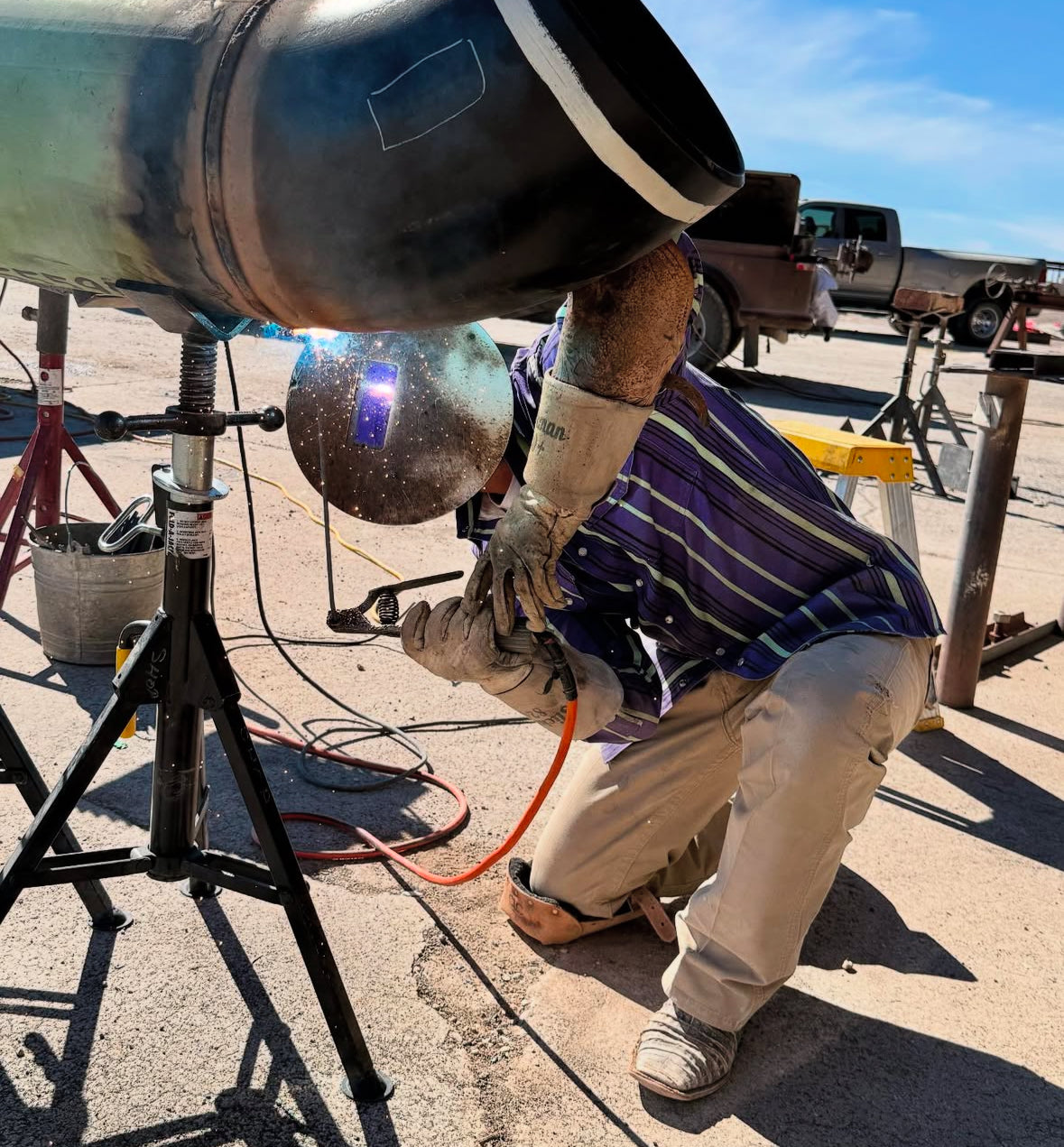 Person welding a large metal object outdoors with a truck in the background.