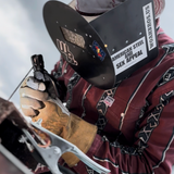 Person wearing a welding helmet with visible branding, holding a welding tool.
