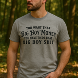 A bearded man wearing a gray “Big Boy Money” T-shirt stands outdoors in a forest setting, sunlight filtering through the trees, highlighting the shirt’s bold black lettering across the chest.