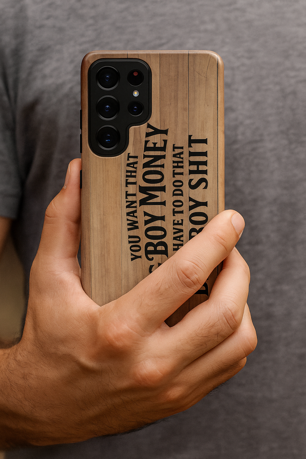 Close-up of a man’s hand holding a Samsung phone with the Big Boy Money tough case, showing the woodgrain design and bold black lettering, “You want that Big Boy Money, you have to do that Big Boy Shit.”