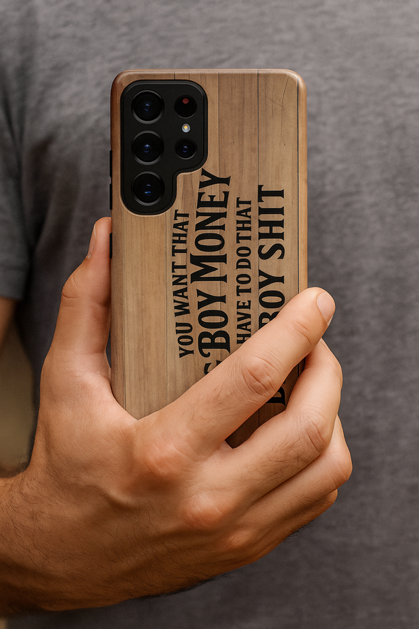 Close-up of a man’s hand holding a Samsung phone with the Big Boy Money tough case, showing the woodgrain design and bold black lettering, “You want that Big Boy Money, you have to do that Big Boy Shit.”